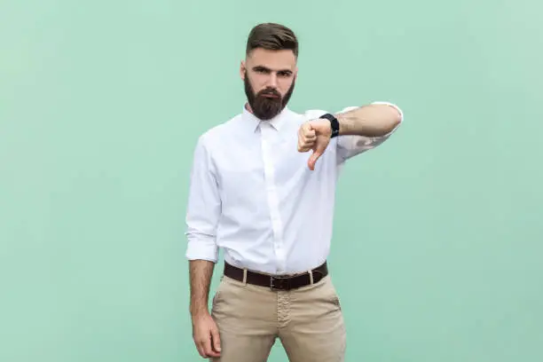 Bearded businessman wearing shirt criticizing bad quality with thumbs down displeased grimace, showing dislike gesture, expressing disapproval. Indoor studio shot isolated on light green background.