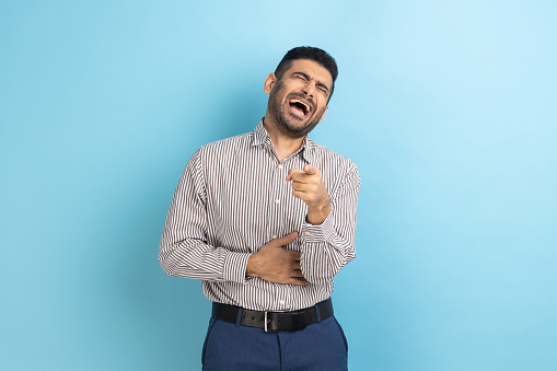 Happy pleased young businessman pointing finger at you and sincerely laughing holding hand on stomach, having fun, wearing striped shirt. Indoor studio shot isolated on blue background.