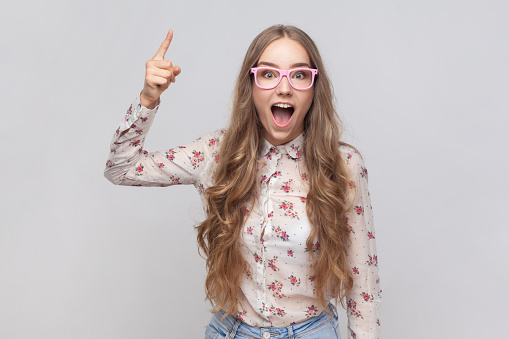 Portrait of inspired woman in glasses with long blond hair pointing finger up and looking amazed about sudden genius idea, got solution. Indoor studio shot isolated on gray background.