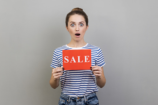 Portrait of surprised woman wearing striped T-shirt holding card with sale inscription, looking at camera with open mouth and big eyes. Indoor studio shot isolated on gray background.