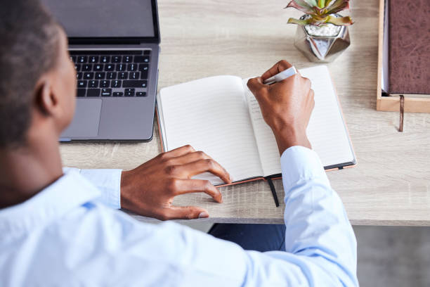 istock Businessman, writing in notebook and planning corporate schedule, calendar and brainstorming idea for lead generation. Black man, write contact list in book and working on advertising strategy goal 1480185111