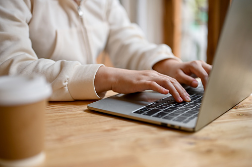 Close-up image of a woman in white hoodie using her laptop, typing on keyboard, browsing internet, remote working at a coffee shop.