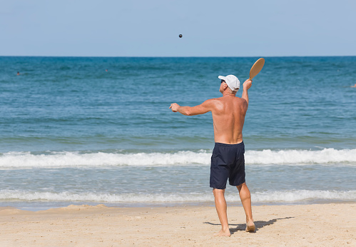 A mature man hits a ball while playing matkot on the beach. Background of the sea. Active life in retirement. Full-height
