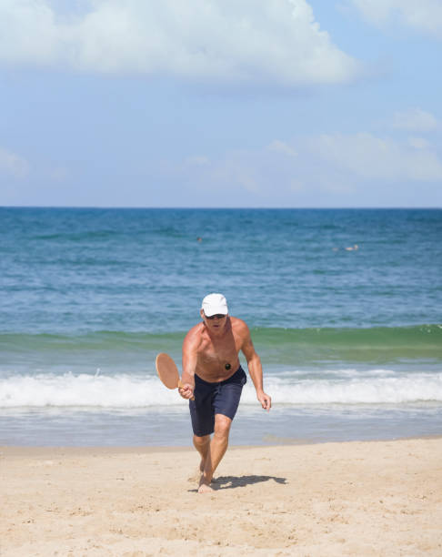 un uomo maturo colpisce una palla mentre gioca a matkot sulla spiaggia - matkot foto e immagini stock