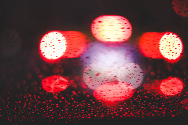 Shiny bright red bokeh lights glowing behind wet glass window with raindrops in dark night time