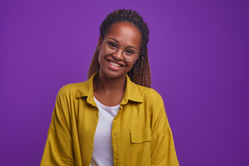 Young smiling attractive African American woman with long curly hair looks at camera wanting to share good mood and positive attitude to what is happening stands on isolated purple background