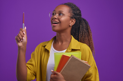 Young inspired beautiful African American woman student with textbooks and workbooks raises pen up to answer question or come up with idea for graduation project stands on purple background