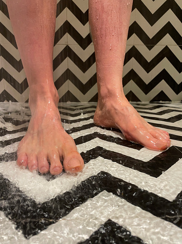 Stock photo showing close-up view of unrecognisable person standing in a puddle in a luxury hotel shower with black and white zig-zag patterned floor and wall tiles.