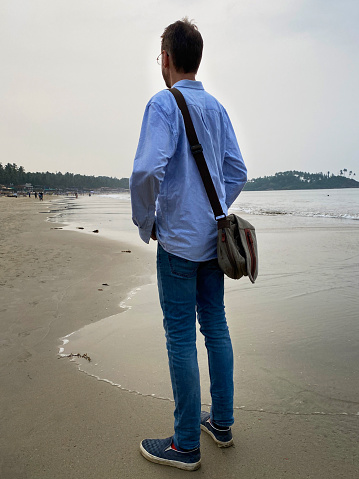 Stock photo showing beach at sunset with caucasian man standing by water's edge of sea.