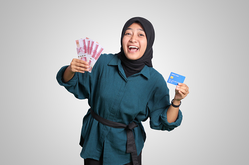 Portrait of cheerful Asian muslim woman with hijab, showing one hundred thousand rupiah while holding a credit card. Financial and savings concept. Isolated image on white background