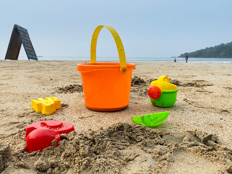 Stock photo showing a collection of multicoloured, plastic beach toys on sandy beach at water's edge of sea. Toys include a bucket, spades, sea animal moulds and a watering can in front of sea backdrop.