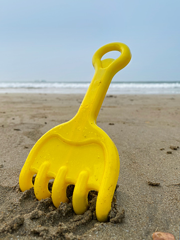 Stock photo showing a brightly coloured, yellow plastic rake beach toy on sandy beach at water's edge of sea.