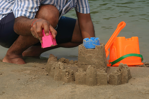 Stock photo showing sandy beach with a sandcastle made with a bucket with a moulded shape, a man is seen making this sandcastle, with plastic children's beach toys.