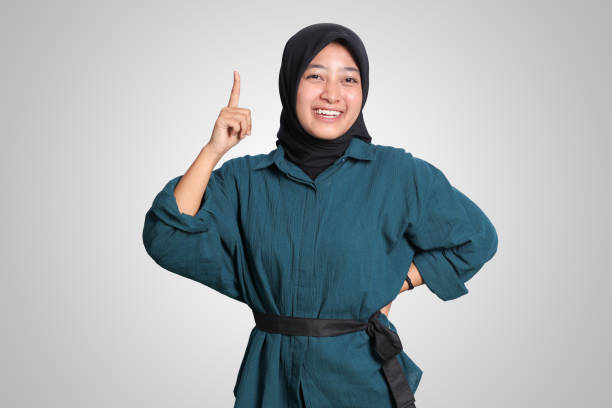 Portrait of excited Asian hijab woman in casual shirt pointing up her forefinger and reminding something to do. Advertising concept. Isolated image on white background stock photo