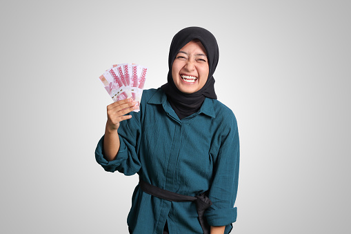 Portrait of excited Asian hijab woman in casual outfit showing one hundred thousand rupiah. Financial and savings concept. Isolated image on white background