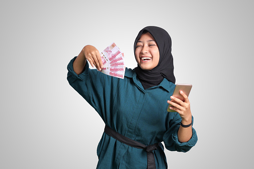 Portrait of excited Asian hijab woman in casual outfit showing one hundred thousand rupiah while holding a mobile phone. Financial and savings concept. Isolated image on white background