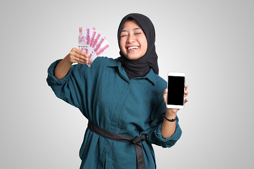 Portrait of excited Asian muslim woman with hijab, showing one hundred thousand rupiah while showing blank screen mockup mobile phone. Financial and savings concept. Isolated image on white background