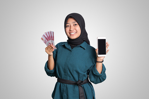 Portrait of excited Asian muslim woman with hijab, showing one hundred thousand rupiah while showing blank screen mockup mobile phone. Financial and savings concept. Isolated image on white background