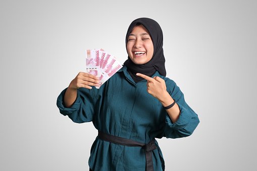 Portrait of excited Asian hijab woman in casual outfit showing and pointing one hundred thousand rupiah. Financial and savings concept. Isolated image on white background