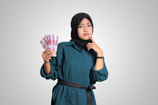 Portrait of confused Asian hijab woman in casual outfit showing one hundred thousand rupiah while thinking with hand on chin. Financial and savings concept. Isolated image on white background