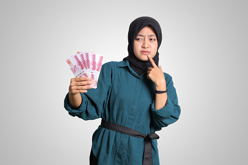 Portrait of confused Asian hijab woman in casual outfit showing one hundred thousand rupiah while thinking with hand on chin. Financial and savings concept. Isolated image on white background