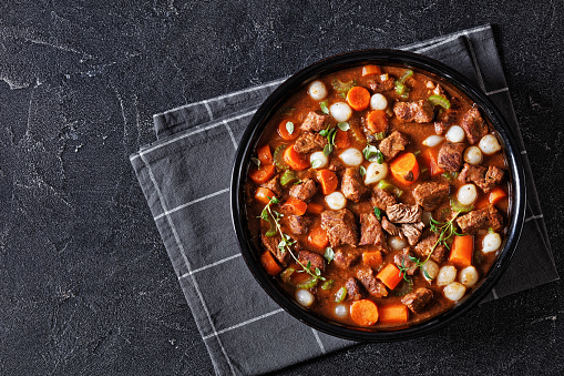 boneless beef short ribs stew with carrots and pearl onions in black bowl on concrete table with napkin, horizontal view from above, flat lay, free space