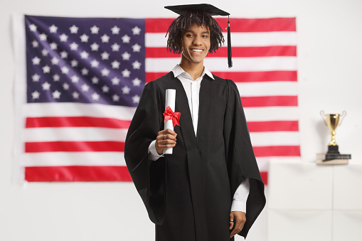 Proud male african american student wearing a graduation gown and holding a diploma in front of the USA flag
