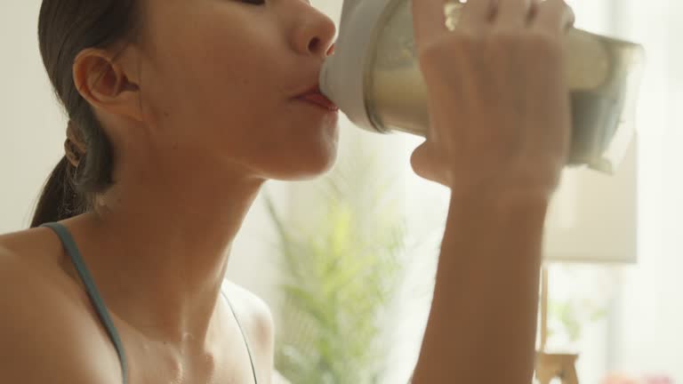 Closeup of young Asian athletic woman drinking protein shake on sofa in living room at home. Diet and healthy food.