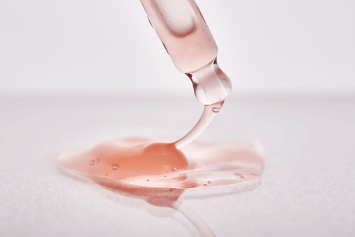 Pipette with a viscous pink cosmetic close-up. The drop stretches from the pipette to the product spilled on the surface.