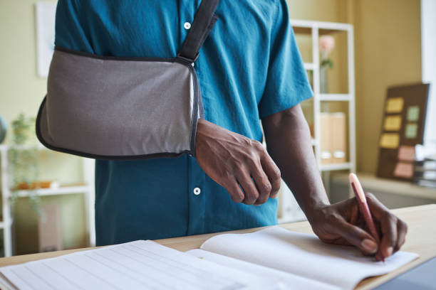 Close up of black man with arm sling working at standing desk in office Close up of black man with arm sling working at standing desk in office and writing in notebook man with arm sling stock pictures, royalty-free photos & images