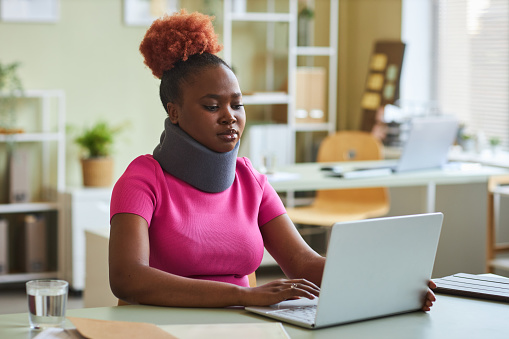 Portrait of black young woman with neck brace working at desk in office and using laptop