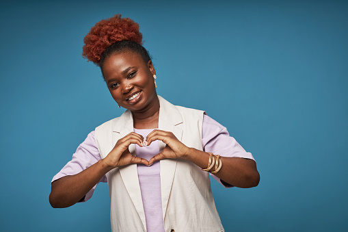 Vibrant waist up portrait of smiling black woman showing heart sign against blue background in studio