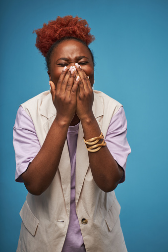Candid portrait of emotional black woman posing against vibrant blue background in studio and covering mouth or screaming