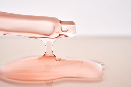 Pipette with a viscous pink cosmetic close-up. The drop stretches from the pipette to the product spilled on the surface.