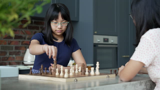 Two young Asian girls playing chess on a dining table, making a move in game of chess. This activity promotes family bonding and allows them to spend quality time together.