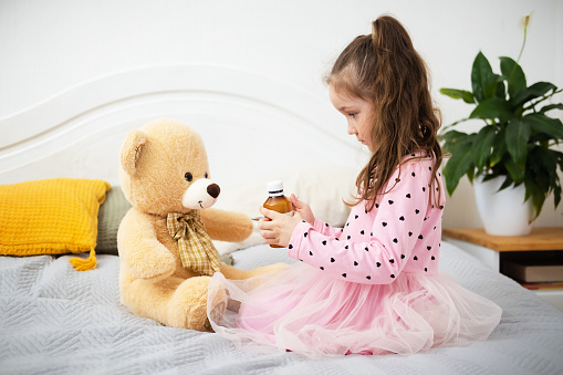 Cute little Caucasian girl is sitting on bed and gives medicine in a spoon from a bottle to teddy bear. Child plays doctor at home