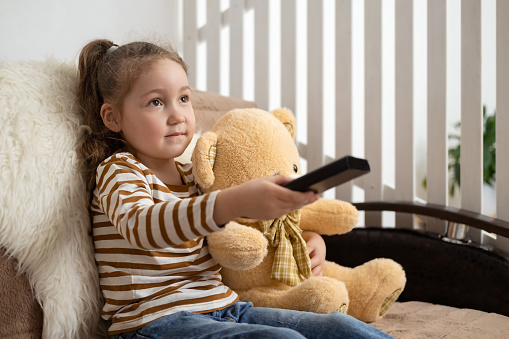 Bored little girl watching tv at home. Child is sitting on sofa with her toy friend teddy bear, holding remote and switching channels, nothing interesting to see. Copy space