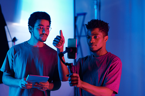 Young African American videomaker using smartphone to make content during teamwork with his colleague in studio