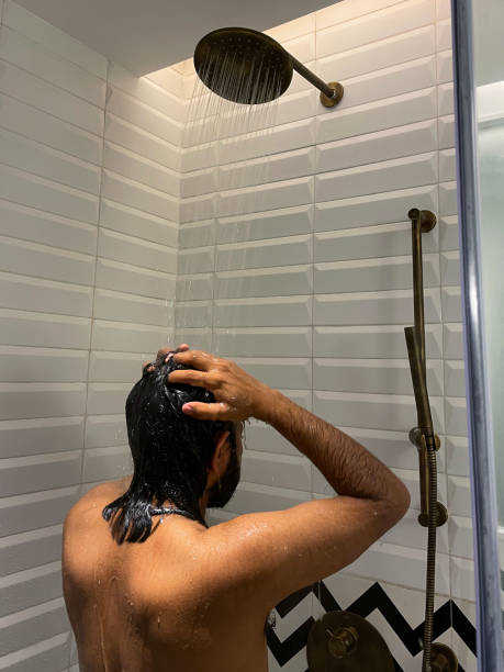 Image of Indian man washing hair in shower cubicle, rinsing soap foam from hair under flowing water, monochrome zig-zag patterned wall tiles, rear view, focus on foreground Stock photo showing close-up view of Indian man rinsing soap suds from his hair under a running shower in luxury, bathroom suite with black and white zig-zag patterned wall tiles. shower men falling water soap sud stock pictures, royalty-free photos & images