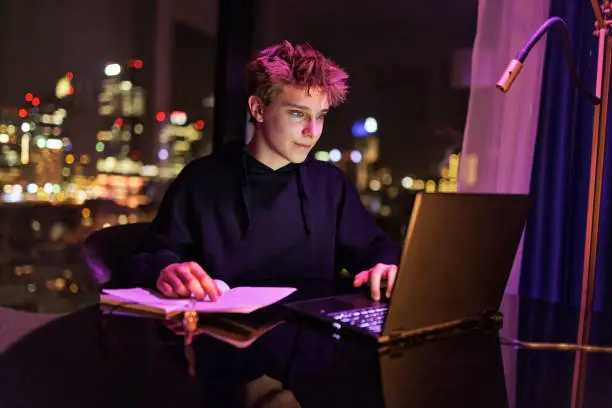 Teenage boy doing homework and studying late at night. Night big city lights in the background. The boy is studying with friends online.
Canon R5