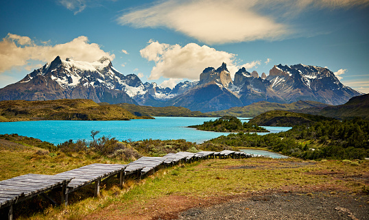 Wooden pathway with snowy Towers of Paine and Paine Horns beneath clouds in background, springtime in Patagonia.