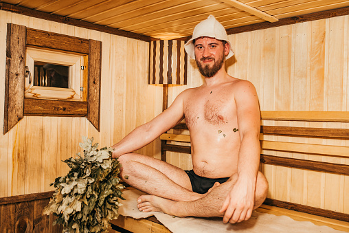 The bath attendant dressed in a special bath clothes is making spa massage to the man using two oak brooms. He is standing on the foreground at Russian steam bath (banya). A caucasian bearded man in a bath cap is lying on his stomach on a wooden bench