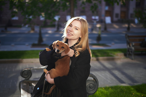 Dachshund puppy. Beautiful blonde young caucasian girl in black jacket standing outside in the city with small puppy in hands looking at camera. Stylish woman with urban background. High quality image