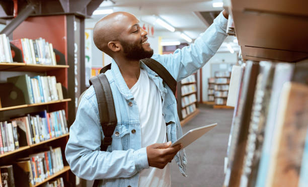 istock Black man, student and library bookshelf  of a university, college and knowledge center. Notebook, happy young person and smile of a male with books for learning and study research info at school 1480262936