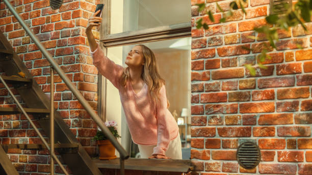 Annoyed Young Female Searching For Mobile Network Connection on Her Smartphone, Reaching Out with a Mobile Phone from an Apartment Window in Urban Neighbourhood.