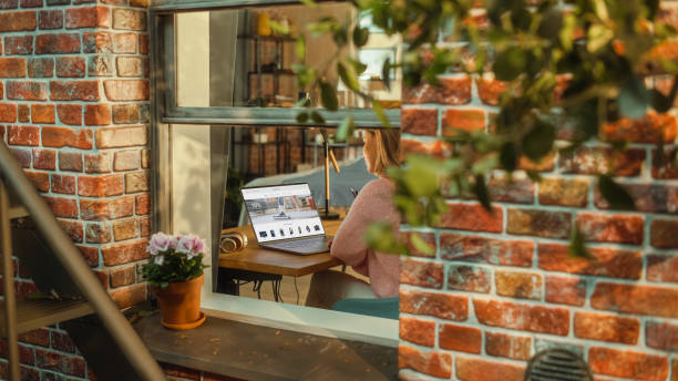 istock Female Working on a Laptop Computer with Online Shopping Website Display while Sitting Behind a Table in her Living Room. Business Manager Working Remotely from Home. View from Outside the Window. 1480264076