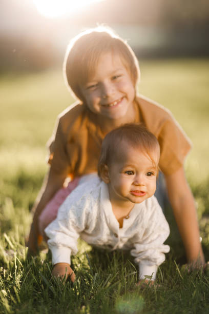 An infant is gnawing a wooden teether toy in the shape of a ring. Drool flows down the hands of the child. Cute sisters are sitting on the floor of the nursery