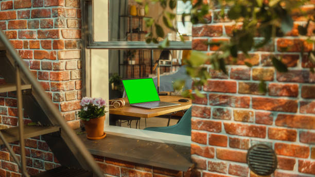 istock Laptop Computer with Green Screen Mock Up Display Standing on a Table in Stylish Living Room. View from Outside the Window of a Brooklyn-Style Brownstone Multi-Storey Building. Work from Home Concept. 1480264190