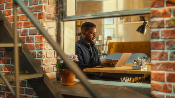 istock Tired African Male Using Laptop at Home. Doing Remote Work During the Day. Frustrated Man Sitting at a Table, Working Long Hours on Computer, Brainstorming Creative Project. Outside the Window Shot. 1480264585