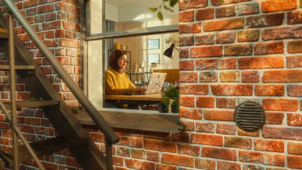 Photo of Young Female in a Hijab Using Laptop Computer, Sitting in Living Room in Her Apartment. Muslim Businesswoman Checking Social Media, Smiling, Answering Emails from Home. Inside Apartment Window View.